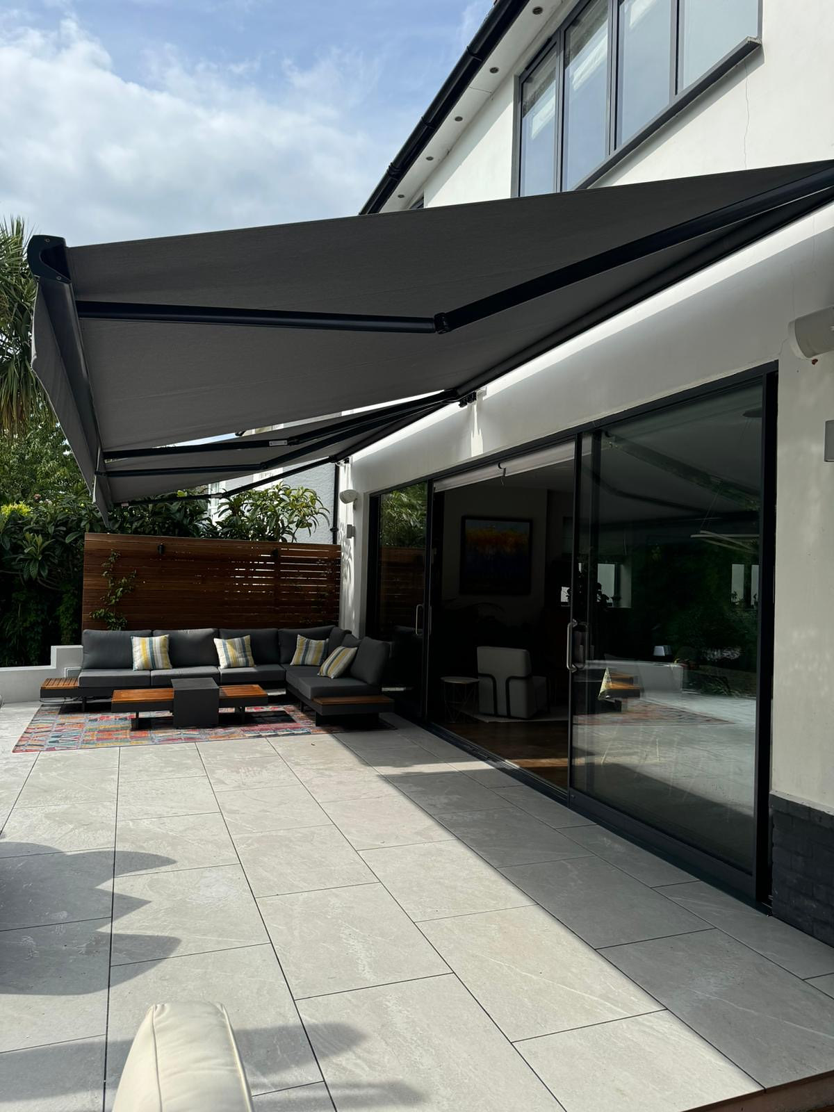 A charcoal folding-arm awning fully extended from a rendered wall, shading an outdoor sofa and dining terrace.