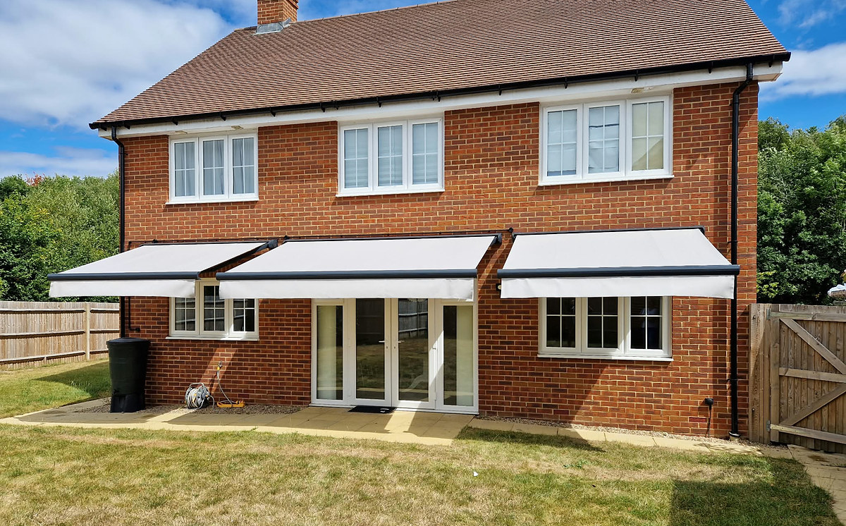 Three linked folding-arm awnings fully extended across the rear of a brick house, shading the patio doors and windows below.