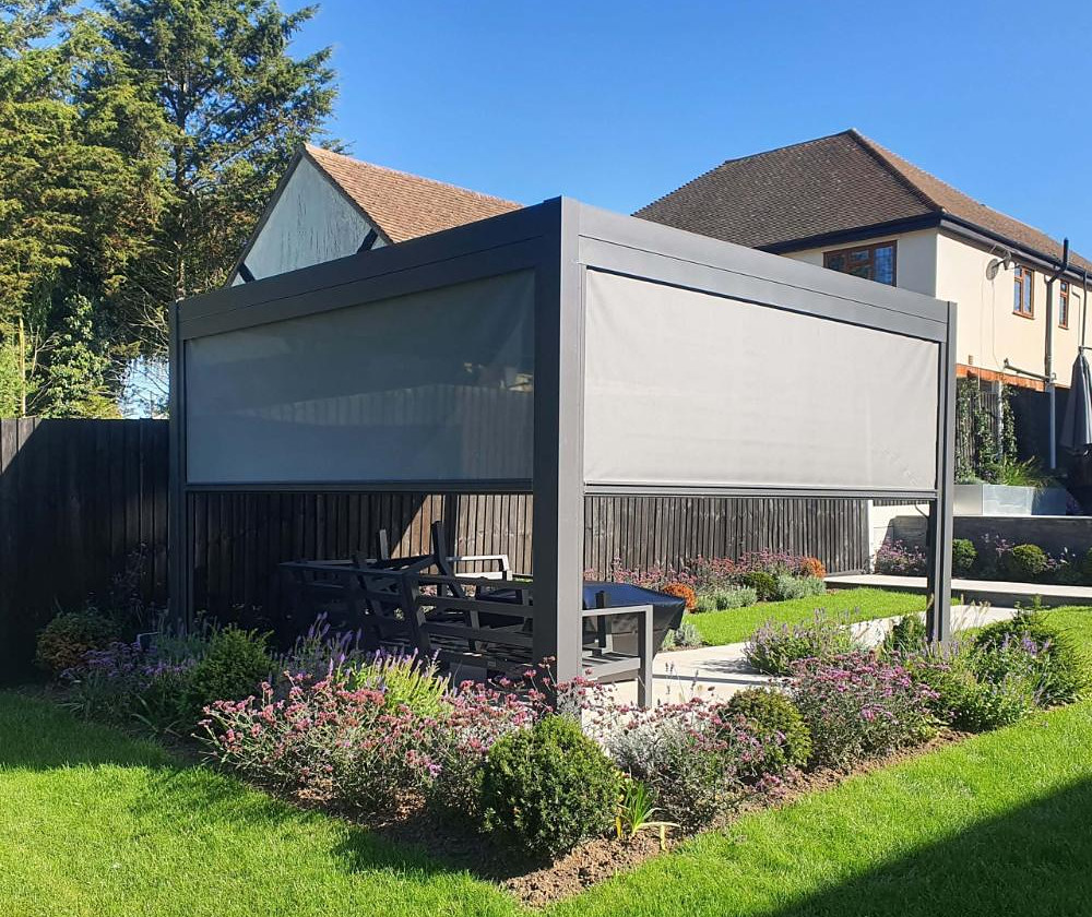 A grey aluminium pergola with zip screens lowered down two sides, standing on a paved terrace in a planted garden.